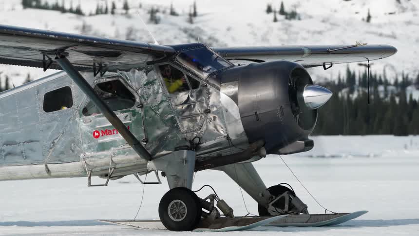 A propeller airplane sits parked in the snow near a forest