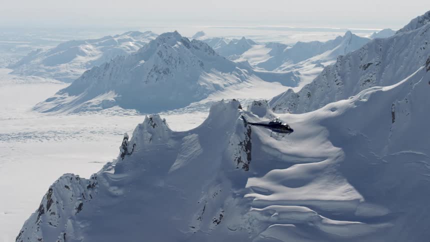 A helicopter rises up and flies over a snow covered ridge on a bright sunny day