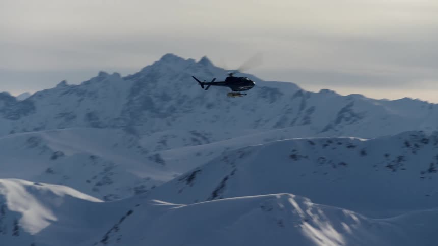 A helicopter flies over snowy mountains under a cloudy sky