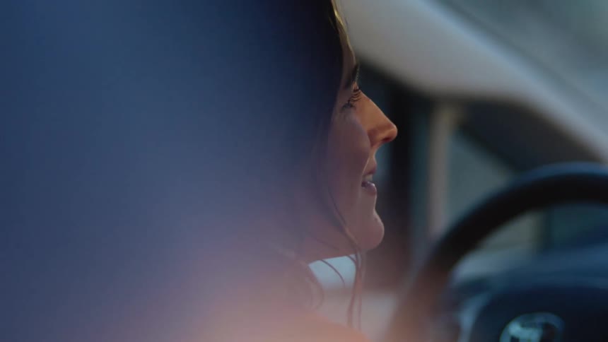A woman sits in her car and smiles and covers her mouth with her hands