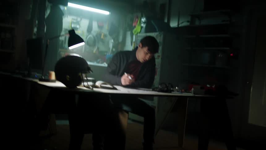 A man sits at a desk in a dark basement and picks up a pen