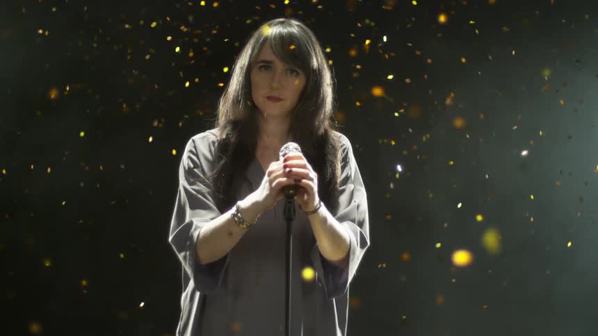 A woman stands still holding a microphone while gold glitter confetti falls and shimmers around her