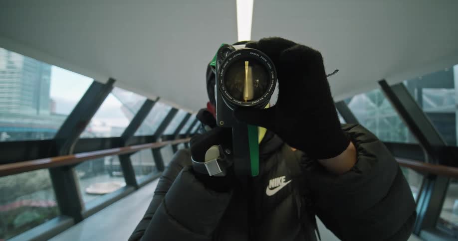 A person holds a camera to their face and adjusts the lens while standing in a window filled tunnel