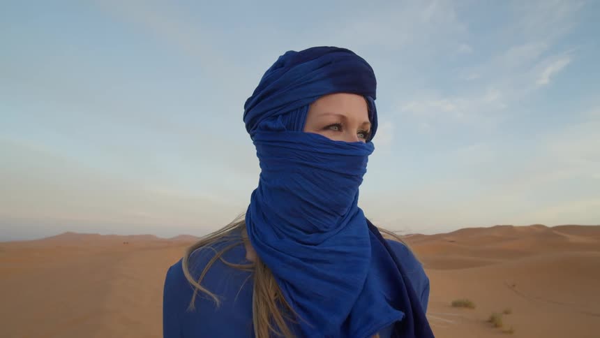 A white woman swathed in blue clothing and a veil stands in the middle of a desert