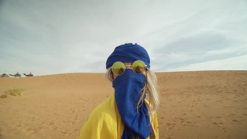 Woman in sunglasses and blue scarf covering head and face in desert with camels in distance