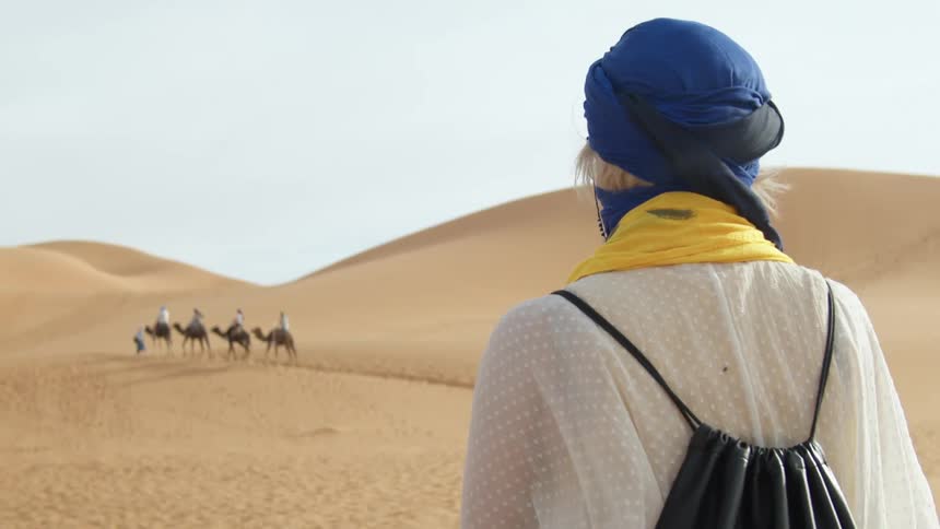 A woman watches people on camels in a desert