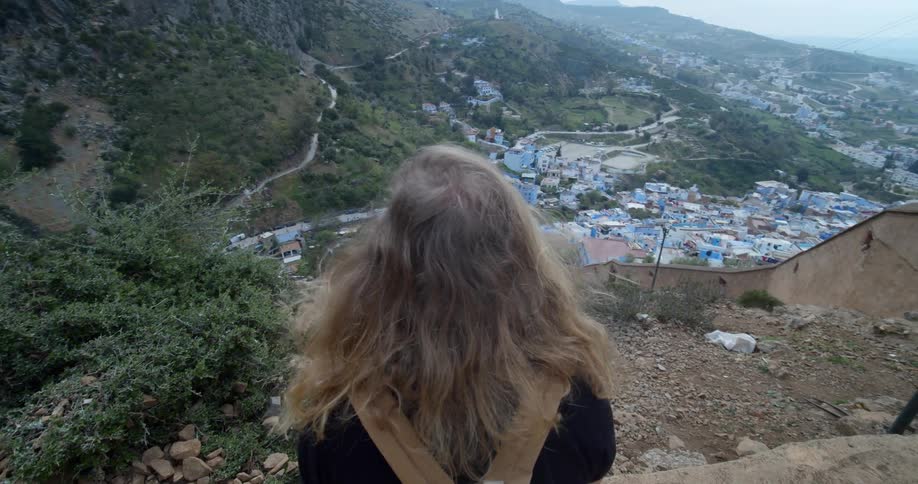 A woman sitting on a cliff overlooking a village and stretching out her arms