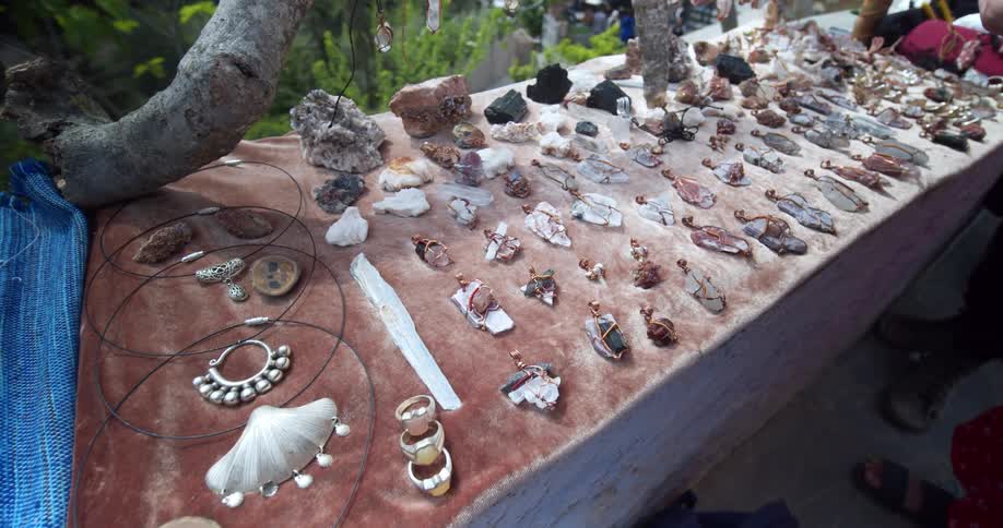 A table of homemade jewelry and trinkets sit on a table outside as people look at them