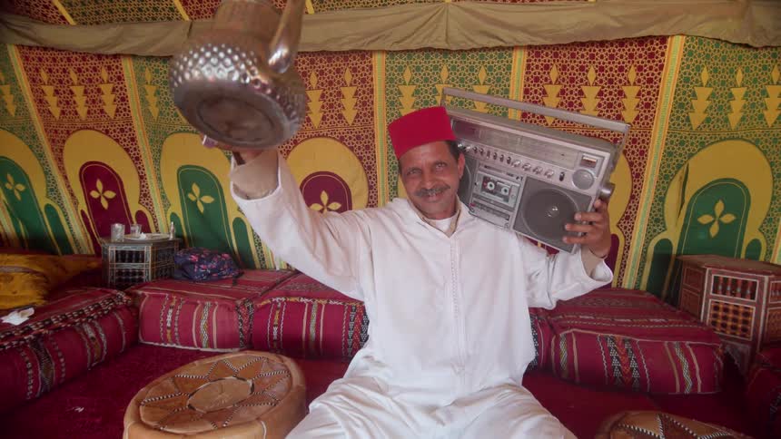 A man holding a radio to his ear pours tea in to cups and gives a thumbs up