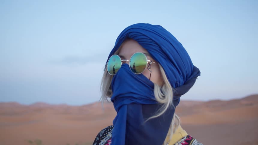 A woman with sunglasses and a scarf on her face standing in the desert with the sky in the background