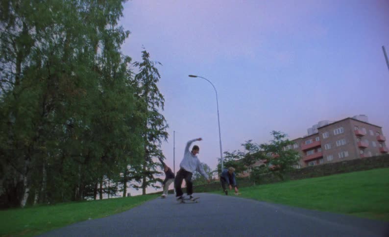 Three white teens skateboard on a concrete path in a city park