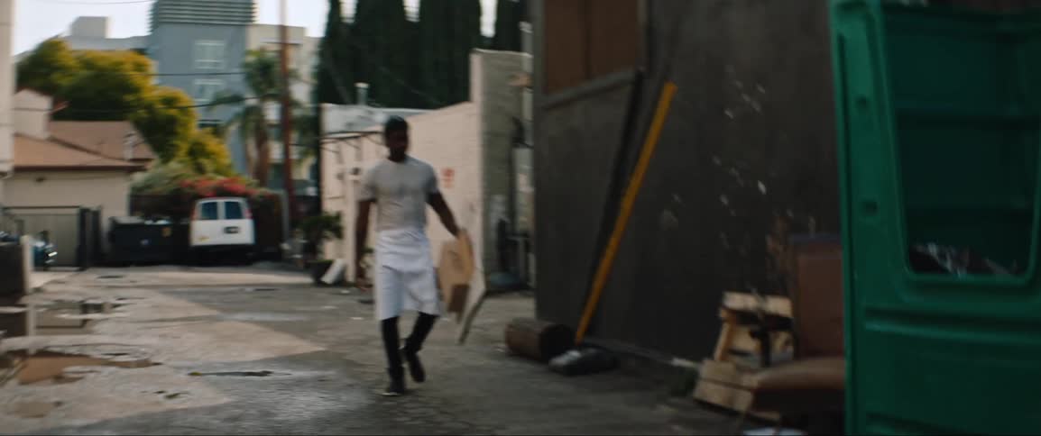 A man in an apron exiting a building into an alleyway to throw cardboard into a large trash bin