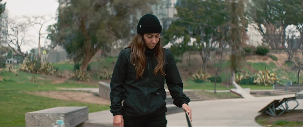 A white woman in black clothing stands in a park and spins her skateboard