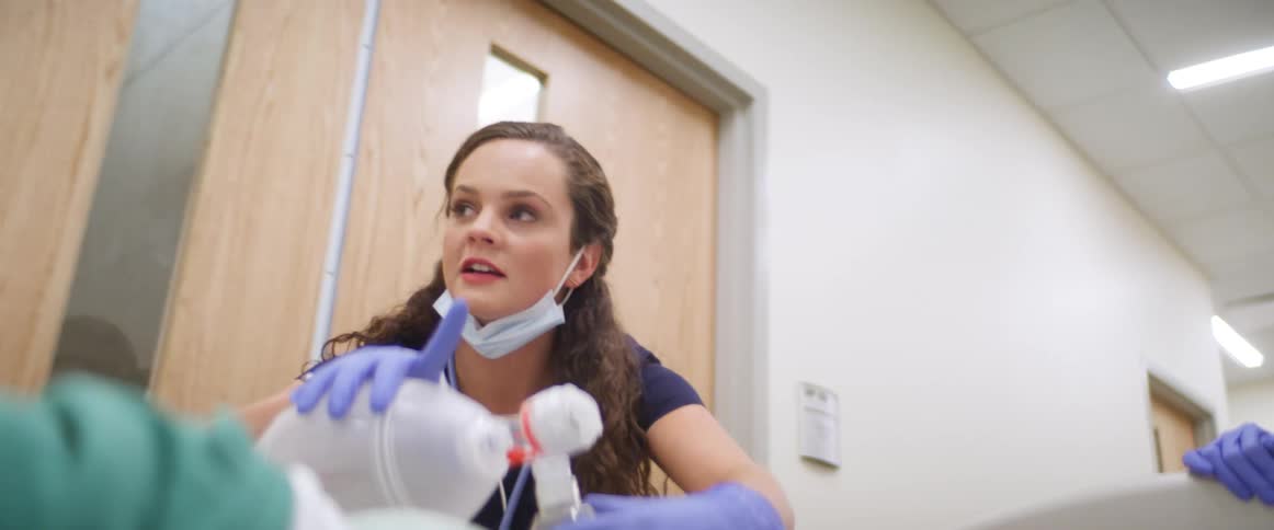 Female nurse wearing a face mask under her chin squeezes ambu bag to resuscitate patient as she walks quickly pushing the patient in a hospital bed through the hallway with a coworker