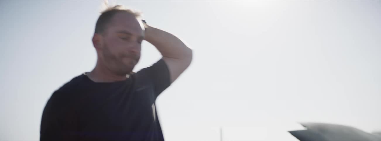 A driver takes off his helmet and walks by a car with smoke coming out of the engine
