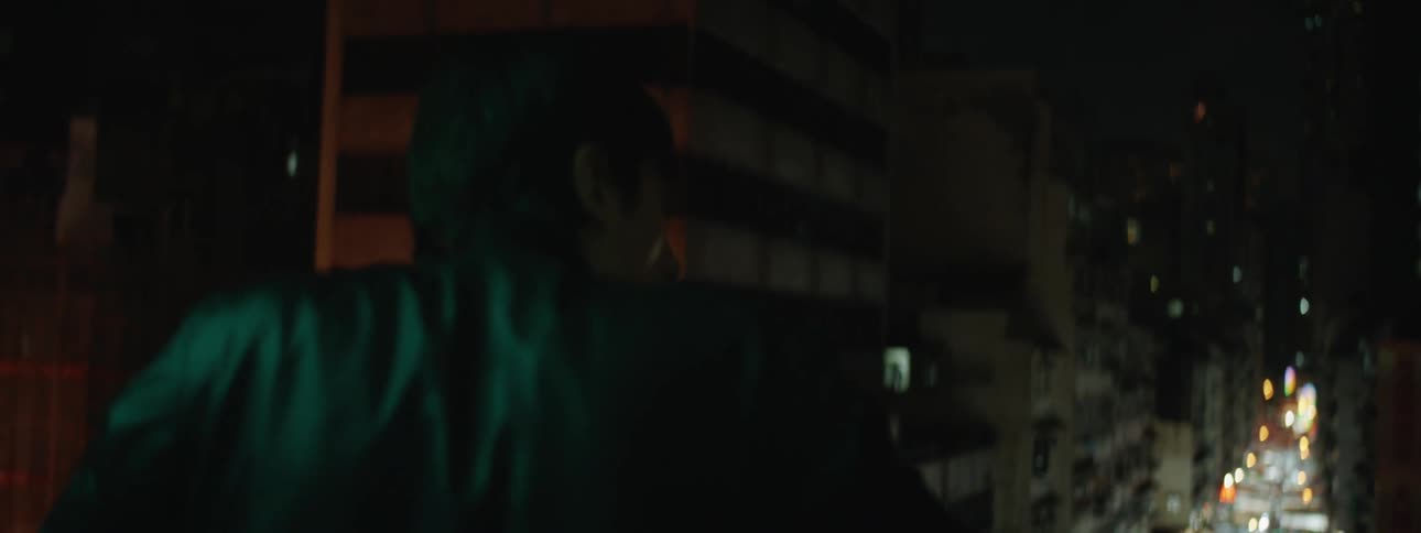 A man in a leather jacket looking out from a balcony over a brightly lit street market at night