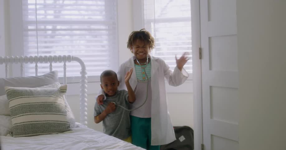 A brother and sister standing in a bedroom smile and wave as they pretend to be doctors