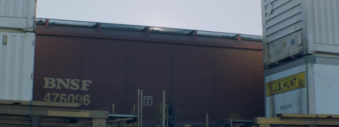 A freight train travels past stacked shipping containers as the sun shines and glares