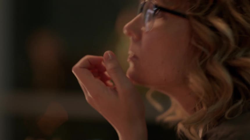A woman repeatedly touches her face and adjusts her glasses while looking at a screen