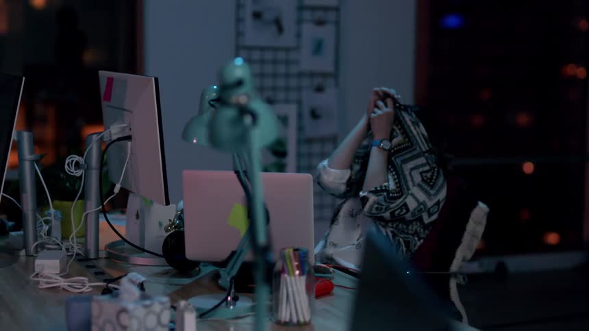 Woman alone in office at night looks around in fear and covers head with sweater