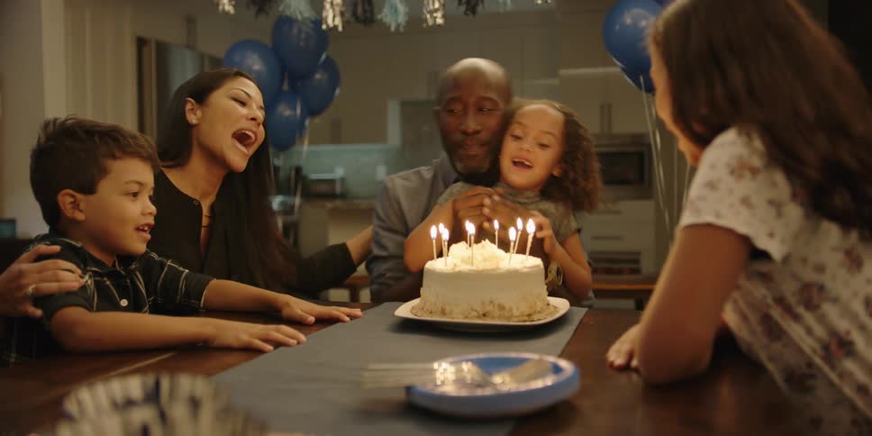 A family sings happy birthday to a young girl seated before a cake with burning candles