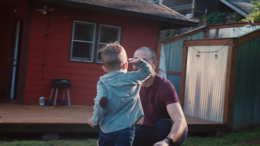 A little boy runs to his dad who lifts him and twirls him in the yard on a bright sunny day