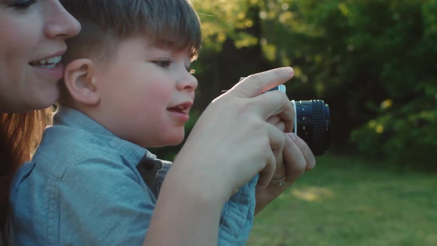 A woman holds a young boy in her lap outside and helps him point and focus a camera