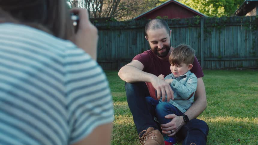 Woman takes a photograph of father and son sitting on the grass in the garden