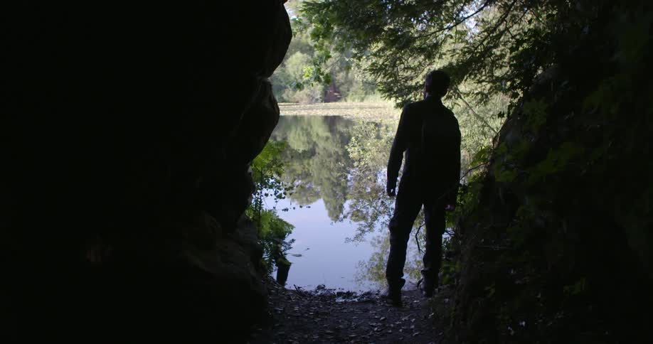 A man walking by a creek with the reflection of trees and the sky coming off of it