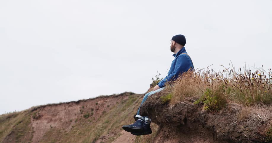 A man sits on the edge of a hill as a bird flies overhead