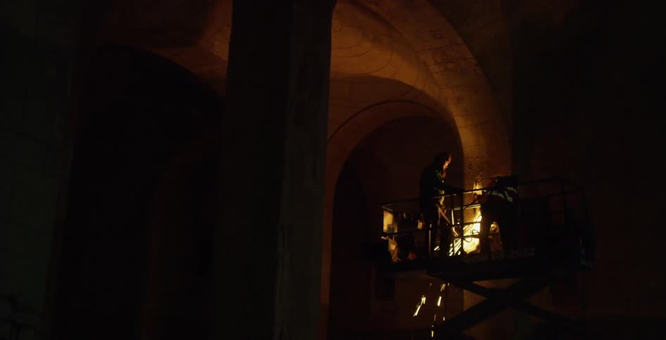Sparks fly and fall from an extendable stand near a high arched ceiling where two construction workers stand