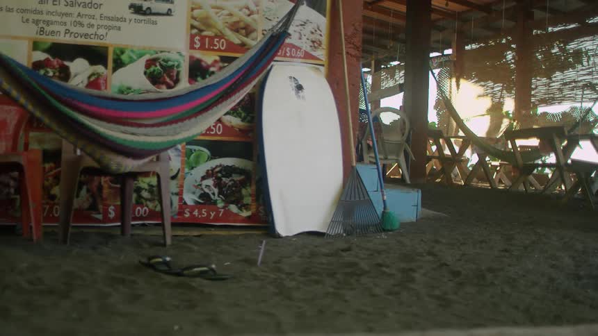 A body board in the sand leans against a menu sign board next to a hammock in an outdoor beach restaurant