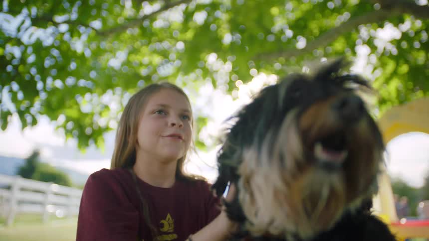 A girl calls out as she sits petting her dog under a tree on a sunny day