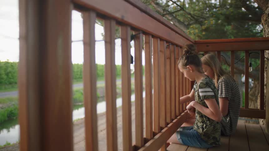 Two young girls sit on the edge of a deck and laugh as someone below on the ground reaches up to them