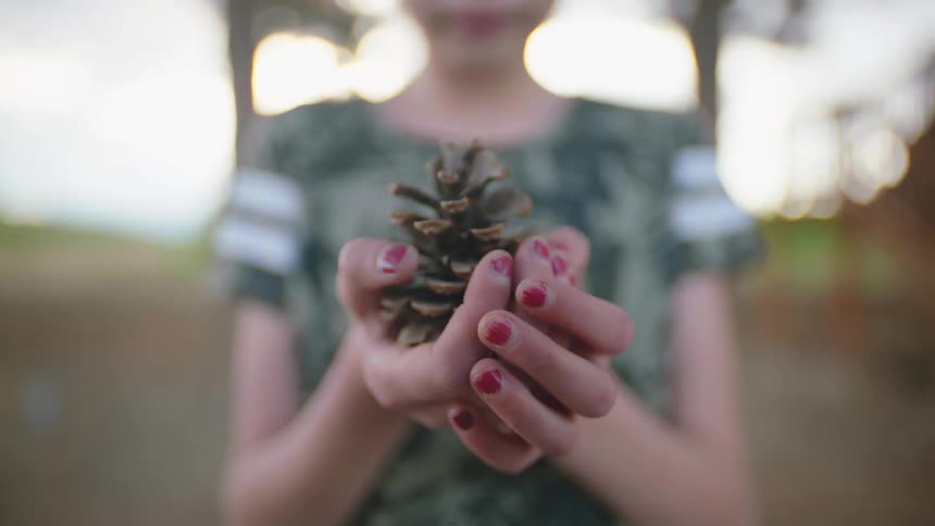 A white girl steps forward carrying a pine cone in her cupped hands