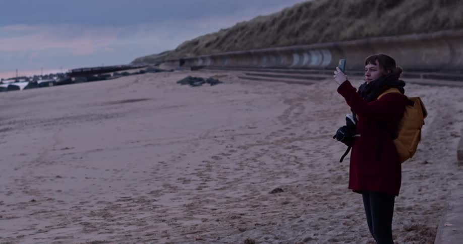 A woman wearing a large coat stands on the beach and points a cell phone towards the ocean water