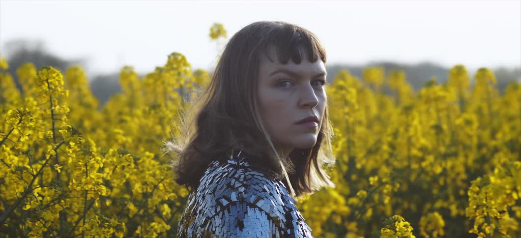 A woman walking through a field of yellow flowers looking at camera