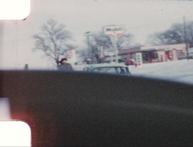 A car driving on a road with snow with buildings and trees in the background