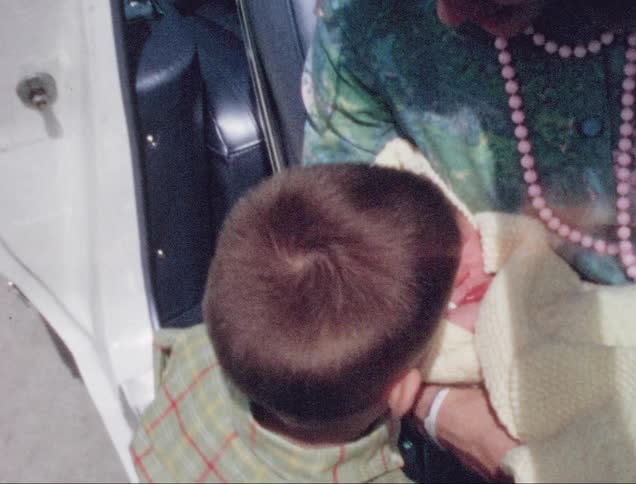 A woman sits in a car with a newborn while a little boy runs over the greet the baby