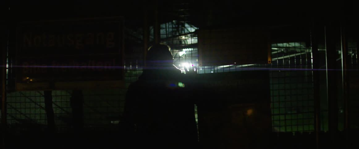 A light shining through a fence onto a person clutching the fence and looking through