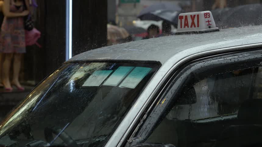 A taxi sits parked on a street with its windshield wipers going during a rain storm