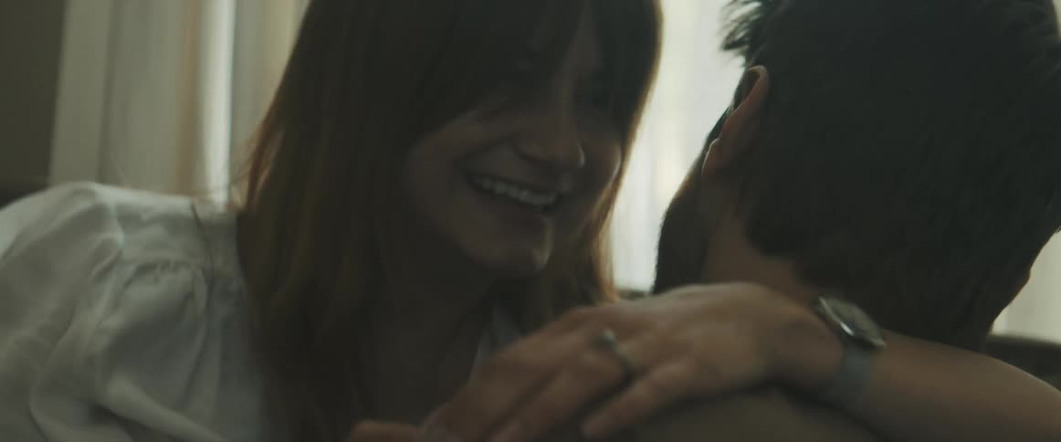 A man and woman hug and laugh on a couch while the woman holds a pregnancy test in her hand