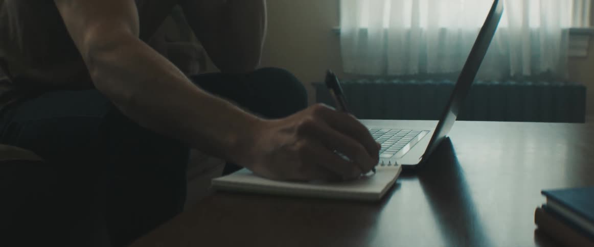 A man sits on a couch talking on the phone while holding a pen to a notepad on a table