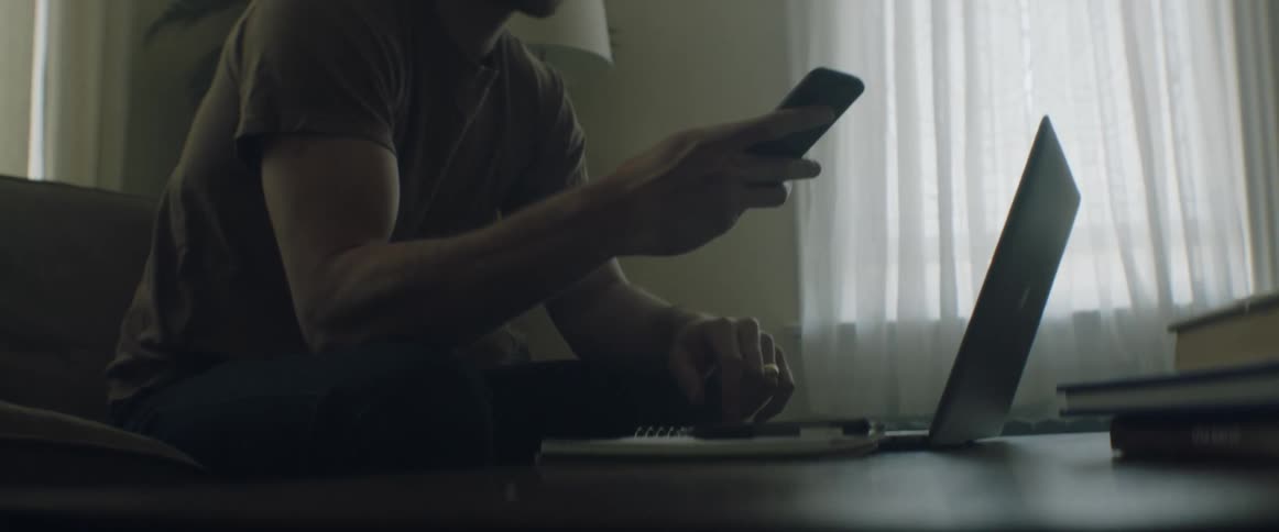 A white man sits in a living room typing on a laptop and then answers a smartphone