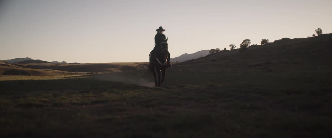 A cowboy rides a horse across a dirt field at sunset