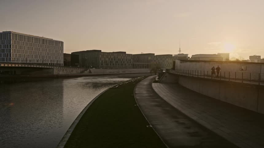 Two people jogging near a canal with buildings and the sky in the background