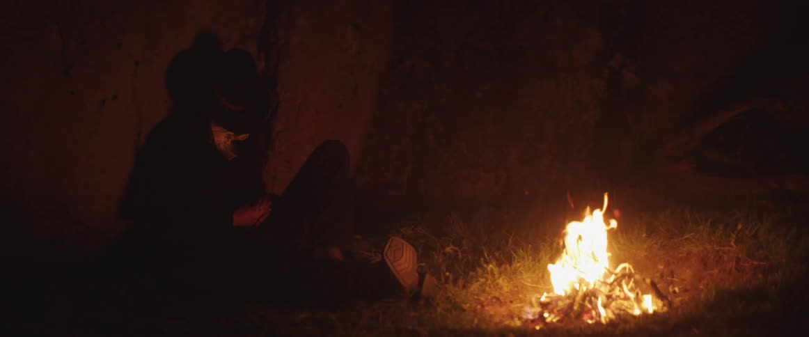 A woman sleeps in front of a burning fire outside