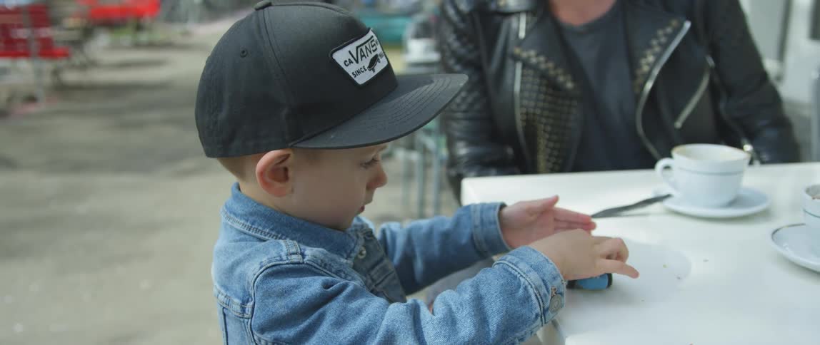 Boy plays with toy car on cafe table as adult sits and watches