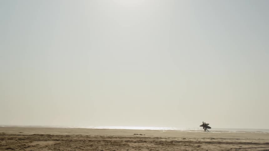 Two surfers walk and jog on the beach with surfboards under their arms