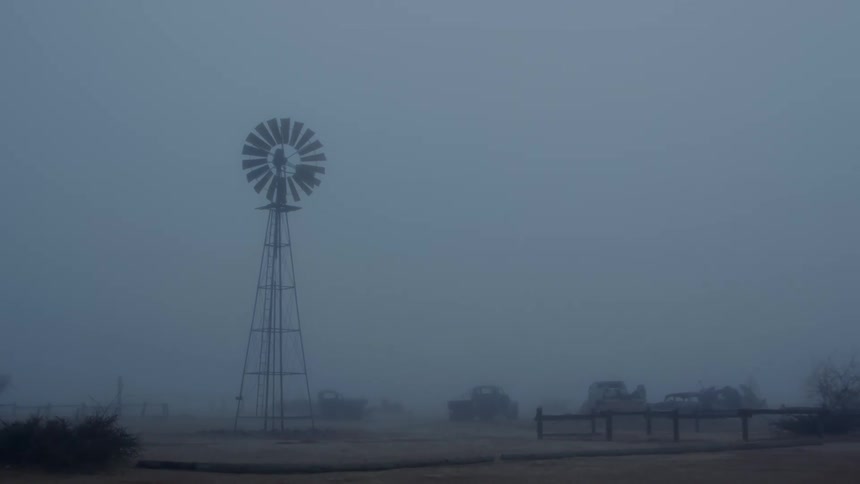 Fog enshrouds vehicles and a windmill in a rural area